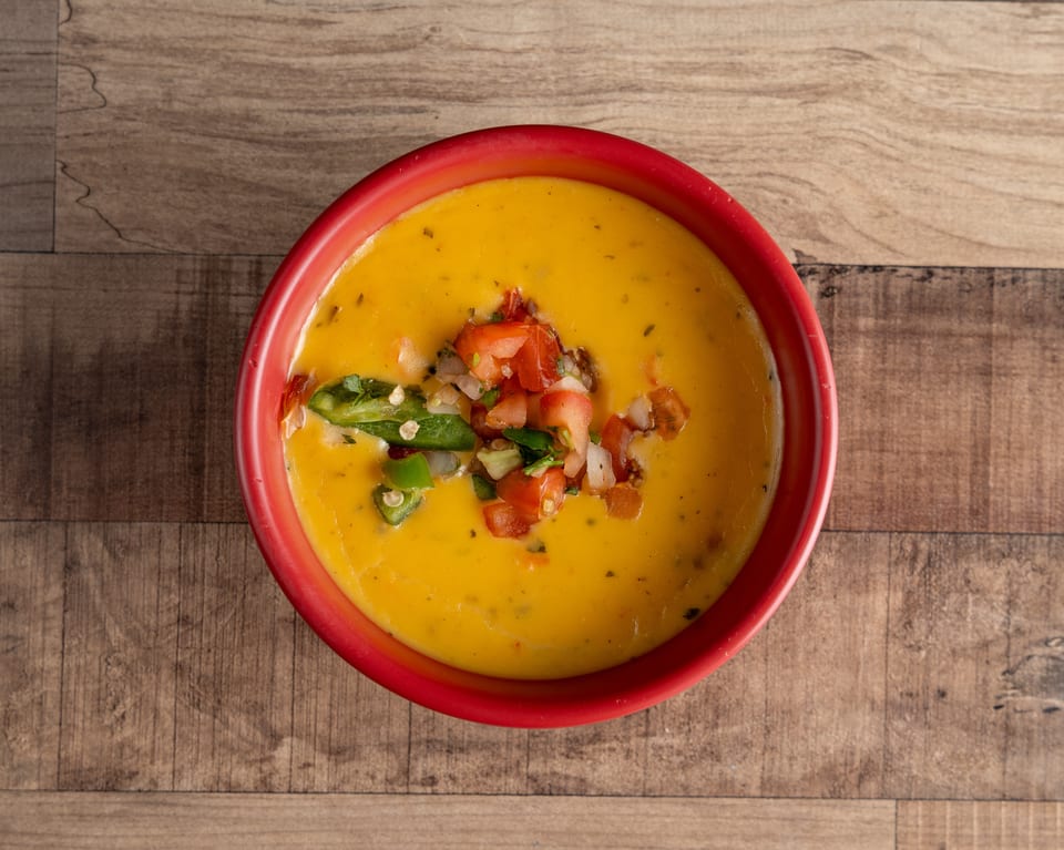 A red bowl of yellow queso dip topped with diced tomatoes, onions, and green peppers.