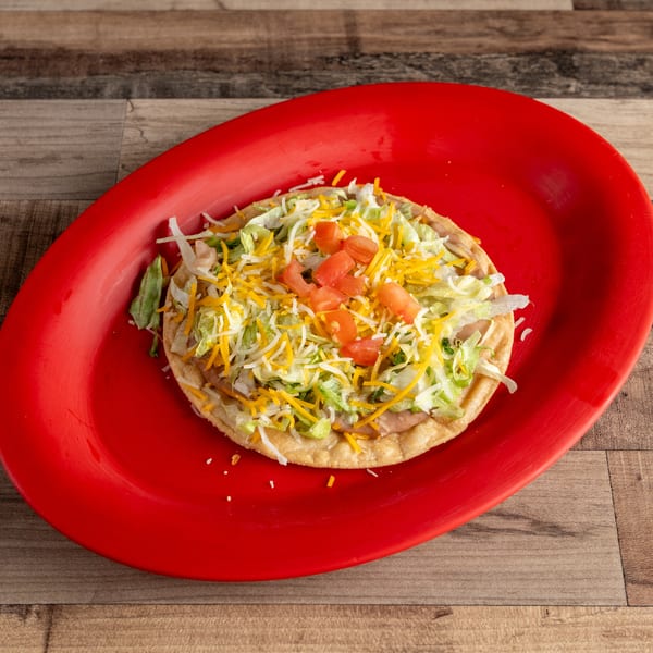 A tostada with refried beans, shredded lettuce, cheese, and diced tomatoes on a red plate.