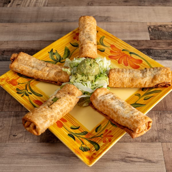 Five golden-brown taquitos arranged around a dollop of guacamole and shredded lettuce on a yellow plate.