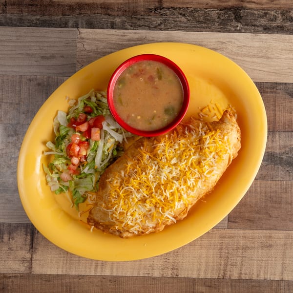 A golden-brown chimichanga topped with shredded cheese, served with a side of lettuce, tomatoes, and a bowl of green chile stew.