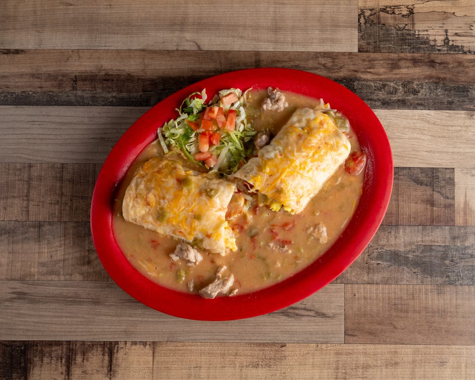 Two cheesy burritos covered in green chile sauce, with a side of lettuce and diced tomatoes.
