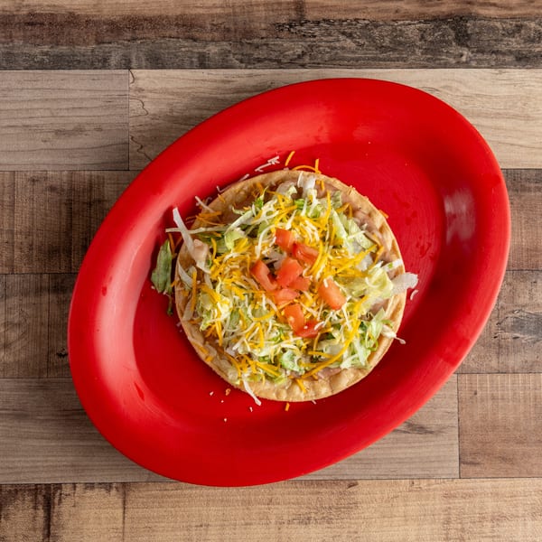 A tostada with refried beans, shredded lettuce, cheese, and diced tomatoes on a red plate.