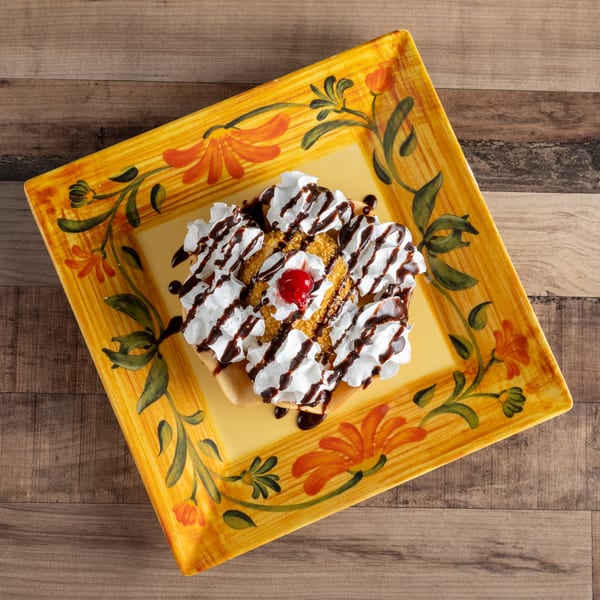 Fried ice cream dessert with whipped cream, chocolate syrup, and a cherry on a decorative plate.
