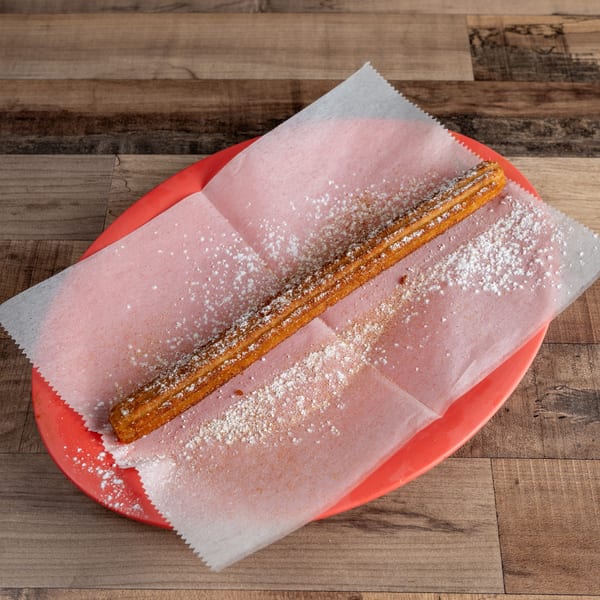 A single churro, dusted with powdered sugar, rests on pink wax paper in a red basket.