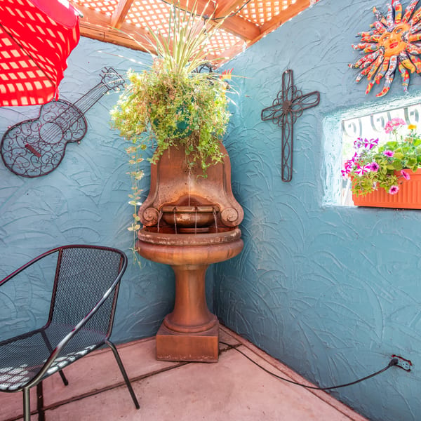 A vibrant patio corner with a fountain, red umbrella, metal chair, and colorful wall decor.