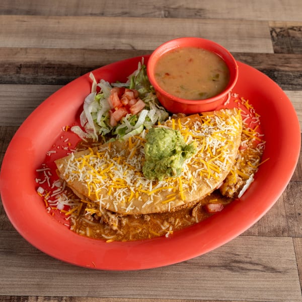 A large fried taco with ground beef, cheese, guacamole, lettuce, and tomato, served with a side of soup.