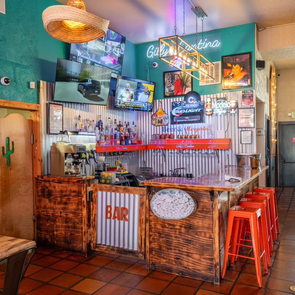 A vibrant bar area with a wooden counter, orange stools, multiple TVs, and neon signs.