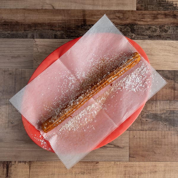 A single churro, dusted with powdered sugar and cinnamon, rests on a red plate with parchment paper.