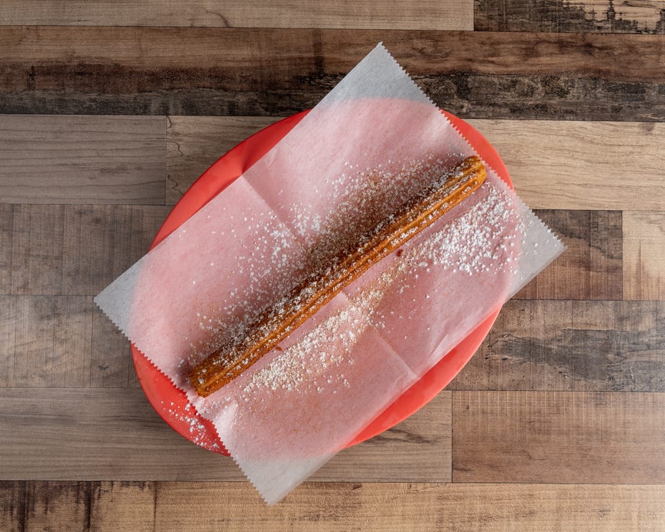 A single churro, dusted with powdered sugar and cinnamon, rests on a red plate with parchment paper.