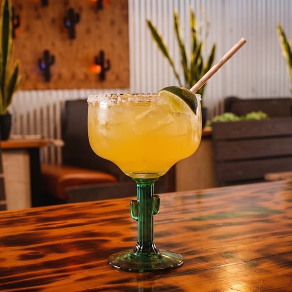 A margarita in a cactus-stemmed glass with a lime and straw sits on a wooden table.