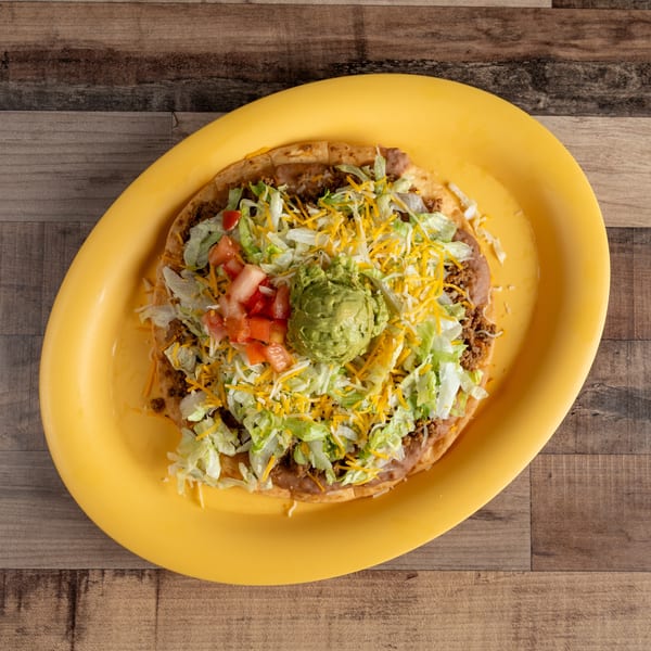 A large Mexican pizza on a yellow plate, topped with ground beef, beans, lettuce, cheese, tomatoes, and guacamole.