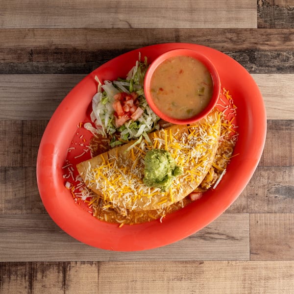A fried quesadilla topped with cheese and guacamole, served with lettuce, tomatoes, and a side of soup.