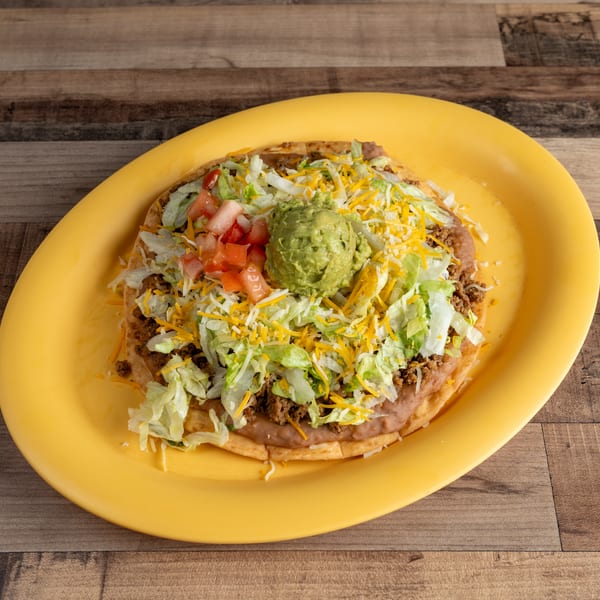 A Mexican pizza on a yellow plate, topped with meat, beans, lettuce, cheese, tomatoes, and guacamole.